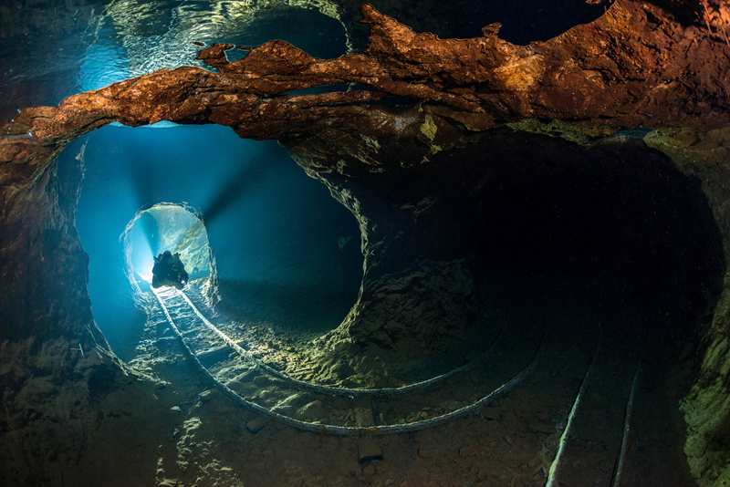 Underwater cave with a diver swimming toward a light source, blue and dark brown tones