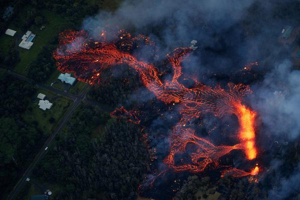 Aerial view of glowing orange lava flows cutting through dark landscape with houses and smoke, dramatic night scene